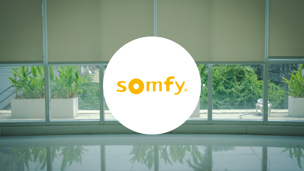 logo Somfy, sur un fond d'une baie voitrée éclairée avec des stores qui remontent